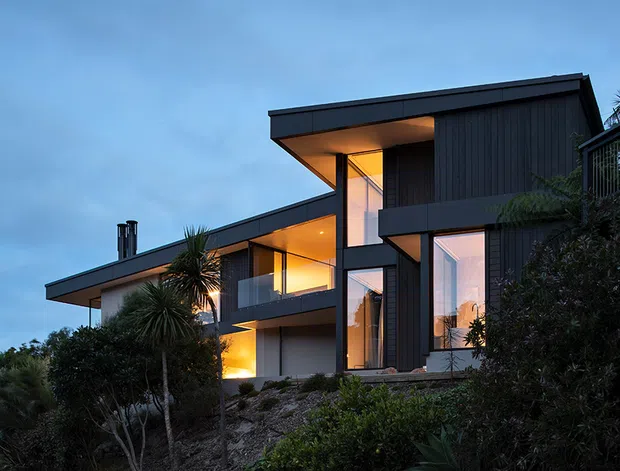 Exterior view of a contemporary multi-level house at dusk, set on a sloped landscaped site. The building features dark vertical cladding, crisp projecting flat roof forms, expansive floor-to-ceiling glazing, illuminated interior spaces, balconies, and strong angular volumes.