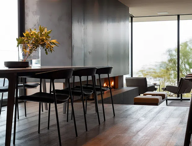 A refined open-plan living and dining interior with a long dark dining table and black chairs in the foreground, opening toward a lounge area and expansive glazing. A linear fireplace is integrated into a dark feature wall, while warm timber flooring, contemporary seating, and a view of trees create a sophisticated, calm atmosphere.