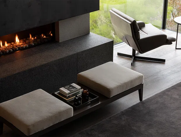A refined contemporary living room with a linear gas fireplace set into a dark textured stone or concrete surround and broad hearth. Two upholstered ottomans sit around a low dark coffee table, while a modern lounge chair faces the fireplace beside full-height corner glazing. The room has a restrained neutral palette, strong geometric forms, and a warm firelit atmosphere.