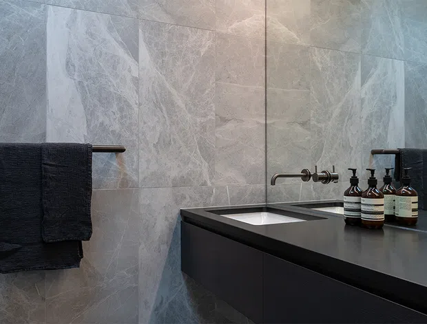 A sophisticated contemporary bathroom vanity area with large-format grey veined stone-look wall tiles, a dark integrated benchtop and floating vanity cabinetry, wall-mounted brushed metal tapware, a large mirror, and dark towels with neatly arranged toiletry bottles. The space has a moody, refined atmosphere and a clean minimalist fit-out.