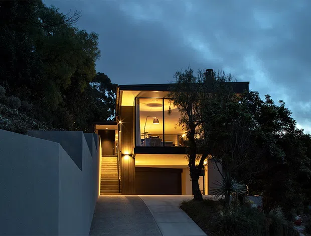 Twilight exterior view of a contemporary multi-level house on a sloping site. The facade combines dark vertical cladding with large illuminated windows, a projecting upper-level living space, an integrated garage, and a concrete driveway and retaining wall. Dense landscaping frames the building, while warm interior lighting creates a strong contrast against the cool blue evening sky.