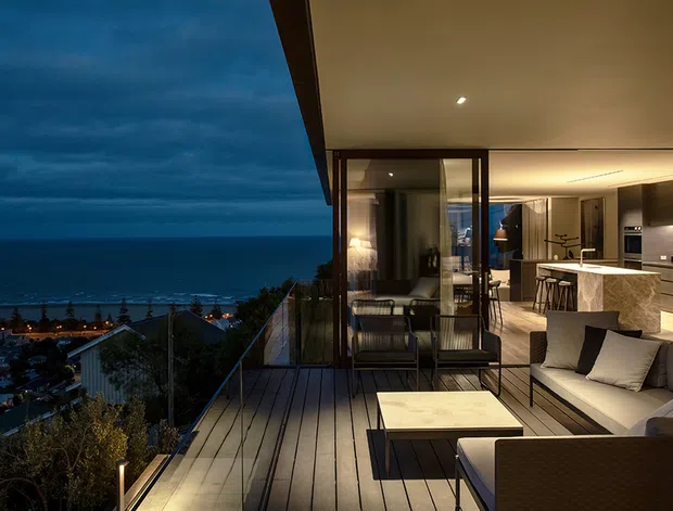 A contemporary coastal residence photographed at dusk, featuring a spacious timber-decked terrace with outdoor lounge seating, glass balustrades, and expansive ocean views. The open-plan interior beyond the glazing includes a refined kitchen with a stone island, dining area, and warm concealed lighting. The scene has a luxurious, calm evening atmosphere with strong indoor-outdoor connection.