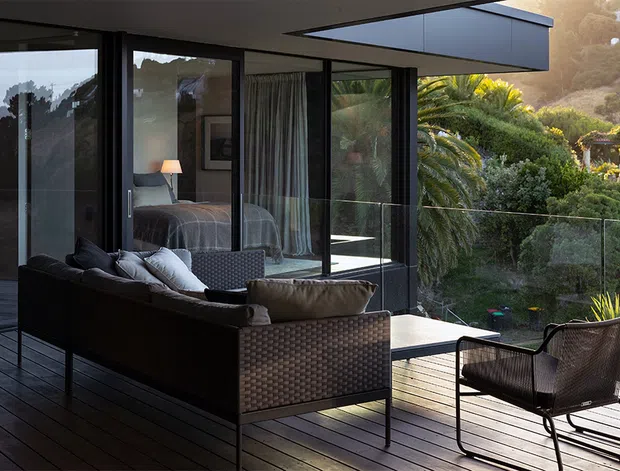 A contemporary indoor-outdoor living area with dark timber decking, black-framed floor-to-ceiling glazing, and a low-profile flat roof. A woven outdoor sofa and armchair surround a small table on the deck, while a bedroom is visible through the glass. Lush landscaped planting fills the view beyond the transparent balustrade.