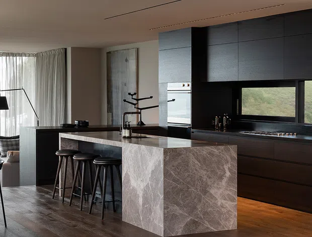 Contemporary open-plan kitchen with a large dark island and waterfall stone end panel, integrated black cabinetry, built-in oven, and a long window above the cooktop. The island includes seating for four on dark stools. Warm timber flooring, recessed ceiling detailing, sheer curtains, and restrained furnishings create a refined, moody yet inviting interior.