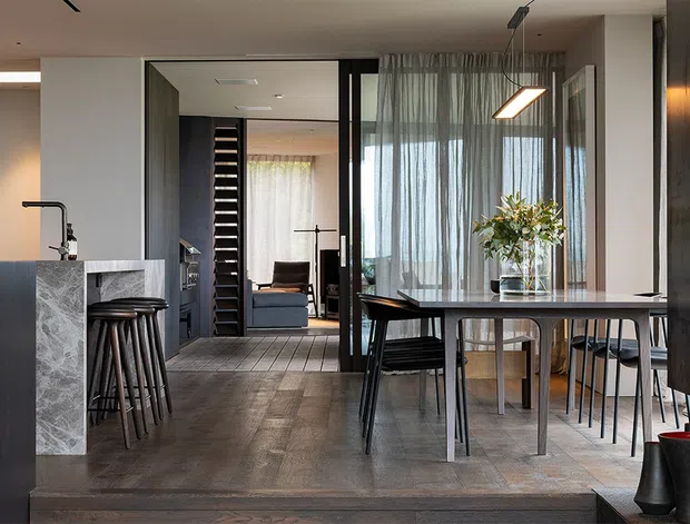 A refined open-plan dining and kitchen interior with a long dining table positioned beside floor-to-ceiling sheer glazing. The space includes a dark kitchen island with a grey stone waterfall end, black bar stools, dark timber flooring, a black-framed opening to a compact living area, and contemporary linear pendant lighting. The palette is muted and sophisticated, combining charcoal, grey, dark timber, and soft natural light.