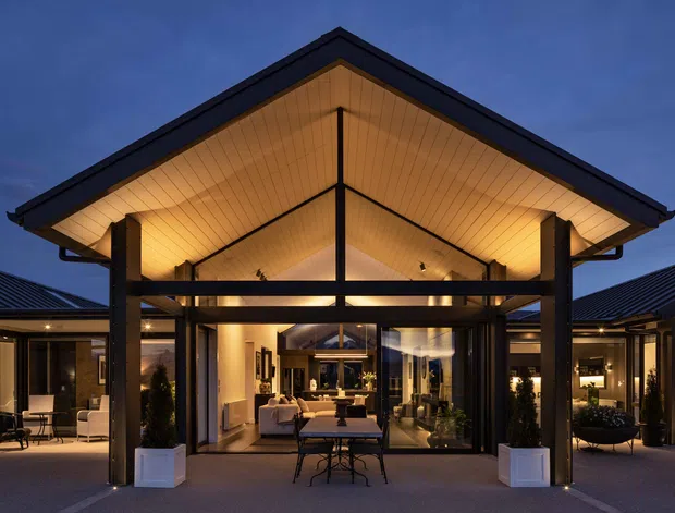 Dusk exterior view of a contemporary single-storey residence centred on a dramatic gabled entry pavilion. The pavilion has a broad pitched roof with warm illuminated timber lining, dark structural posts and beams, and extensive floor-to-ceiling glazing opening onto a furnished outdoor entertaining area. Low wings extend to both sides with dark metal roofs and glazed interiors visible beyond the central opening.