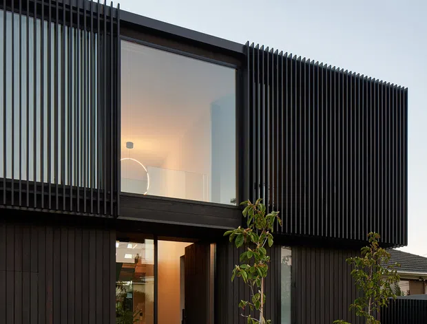 Contemporary two-storey residential facade clad in dark vertical timber-like boards, with prominent black vertical fins screening the upper-level glazing. A recessed entry sits beneath the projecting upper volume, opening onto a pale concrete path through landscaped planting. Warm interior lighting contrasts with the dark exterior, while a neighbouring roofline is visible at right.