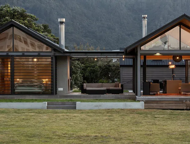 Exterior view of a modern multi-volume house set against a wooded mountain backdrop. The composition features two pitched-gable wings connected by a lower central section, extensive floor-to-ceiling glazing, dark metal framing, timber-toned interiors, and covered outdoor living areas. Warm interior lighting contrasts with the cool, subdued landscape and lawn.
