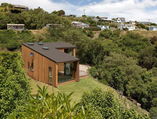 Exterior view of a compact contemporary timber-clad dwelling set into a densely vegetated, sloping site. The single-storey structure has a dark low-pitched roof with several skylights, extensive glazing at the corner, a small timber deck, and a landscaped lawn. Other homes are visible on the hillside in the background.