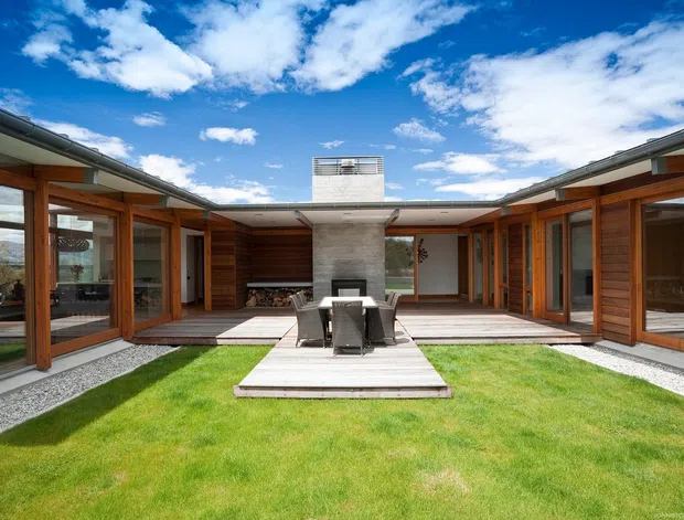Single-storey contemporary courtyard residence viewed from the landscaped central lawn. Two wings with extensive floor-to-ceiling glazing and warm timber-framed doors surround a paved outdoor dining terrace, while a central concrete chimney or feature wall anchors the composition. The building has low-pitched rooflines, crisp horizontal forms, gravel edging, and a bright open-sky setting.