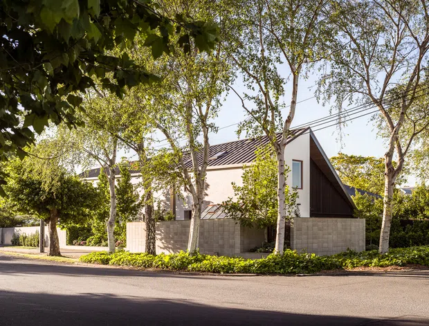 Street-facing exterior view of a contemporary residential house set behind a low concrete block wall and landscaped verge. The home has a steep gabled roof with dark metal roofing, pale rendered walls, a dark vertical timber-clad gable end, and carefully maintained mature street trees and hedges.