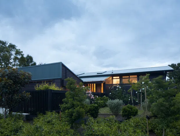 Exterior view of a low-rise contemporary residential house set within a lush landscaped garden. The building has dark horizontal cladding, a corrugated metal gabled roof, broad timber-framed glazed openings, and warm interior lighting visible through the windows. Dense planting and fencing partially screen the facade.