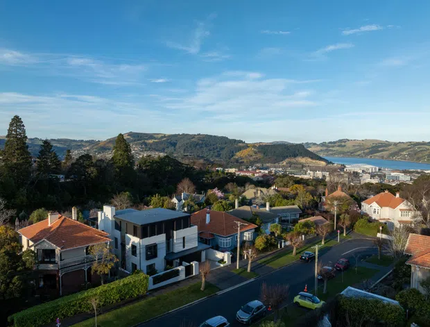 Elevated exterior view over a suburban residential neighbourhood, with detached homes, tree-lined streets, modern and traditional houses, and a broad coastal harbour or inlet visible beyond the hills. The scene is photographed in clear daylight with a wide landscape setting.
