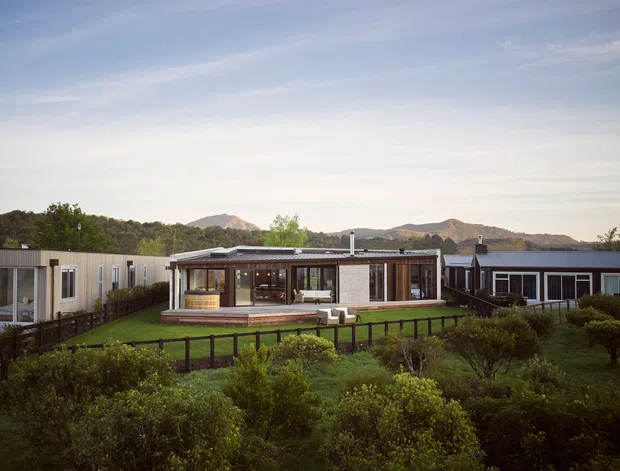 A low-rise contemporary residential complex set within landscaped gardens, viewed from the exterior. The central pavilion has extensive glazing, timber-framed openings, a low-pitched roof and a raised timber deck, with additional connected or adjacent flat-roofed buildings on either side. Mountains and mature vegetation form the background.
