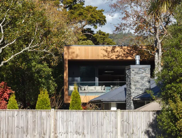 Exterior view of a contemporary two-storey dwelling set among mature greenery. The building features a warm timber-clad upper volume, extensive dark glazing, a projecting balcony or living area, and a prominent rough-stone chimney with a dark pitched roof element. A timber boundary fence and landscaped planting occupy the foreground.
