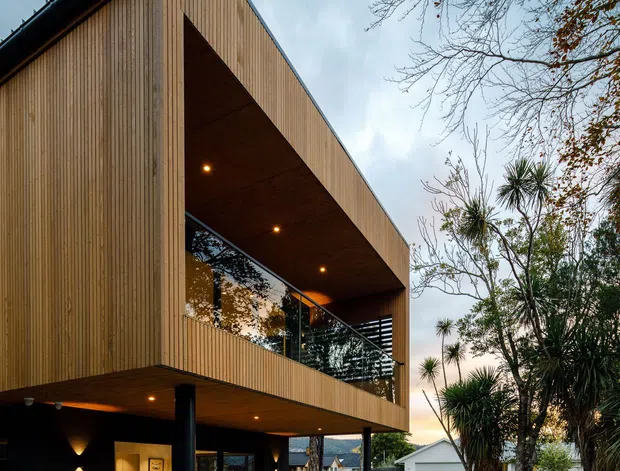 Exterior architectural photograph of a contemporary multi-level home with a sharply folded gable-like roof form, extensive vertical timber cladding, deep cantilevered upper-level overhangs, dark structural columns, and a covered outdoor dining area. Large glazed openings reveal interior spaces, while stone boundary walls, paving, and established planting surround the house.
