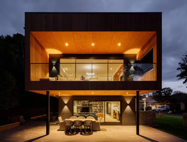 Twilight exterior view of a contemporary two-storey house with a strongly framed, cantilevered upper level. Warm recessed lighting illuminates a timber-lined upper balcony or living area behind expansive glazing, while the covered ground-level outdoor entertaining area includes a dining table and chairs. Dark vertical exterior cladding, slender structural posts, glass balustrades, and a landscaped suburban setting are visible.