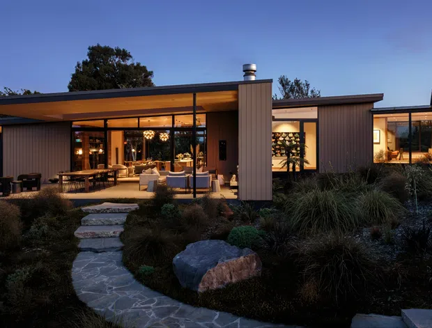 A low, single-storey contemporary home photographed at dusk, with broad glazed openings, dark slender framing, and projecting flat roof forms. Warm interior lighting reveals an open living and dining area, while a stone stepping path crosses a landscaped garden of rocks and ornamental grasses toward the covered outdoor terrace.