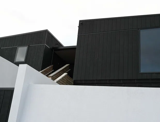 Exterior view of a contemporary multi-level building with dark vertical cladding, large dark-framed windows, and crisp white rendered walls. An exposed timber staircase connects the offset upper volumes, creating a strong geometric, minimalist facade composition.