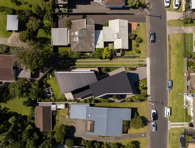 Aerial top-down view of a suburban residential neighbourhood, showing detached homes with varied pitched and gabled roofs, driveways, narrow front gardens, mature trees, and a paved residential street with parked cars.