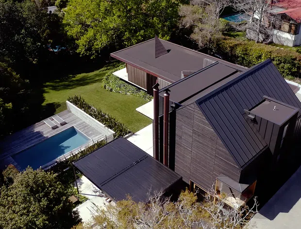 Aerial view of a contemporary residential property set within a landscaped suburban site. The dark metal-clad main house has multiple pitched roof forms, vertical timber-toned wall cladding, and prominent twin chimney stacks. A separate low-roof wing and covered outdoor structure surround a lawn and rectangular swimming pool, with a curved driveway along the right side.
