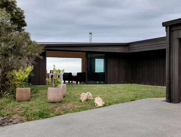 Exterior view of a low, dark-clad modern house with a broad flat or very low-pitched roofline, deep recessed glazing, and a central indoor-outdoor opening. A paved driveway or forecourt occupies the foreground, with lawn, large planters, and landscaped planting around the building. Dark vertical timber-like cladding contrasts with expansive clear glazing and an overcast sky.