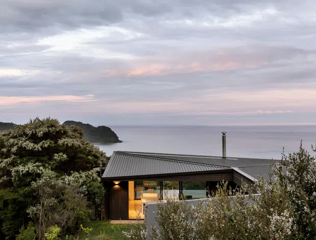 A modern coastal house viewed from the surrounding landscaped site, with dark vertical cladding, a low-pitched corrugated metal roof, broad glazing, and warm interior light. The home overlooks a calm expansive body of water and a wooded headland beneath a soft overcast evening sky. Dense trees and shrubs partially screen the lower facade.