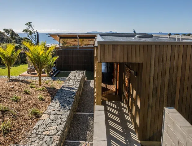 Exterior view of a low-profile contemporary coastal house, with dark vertical timber cladding, a flat metal roof, sheltered outdoor circulation, and textured stone retaining walls. The landscaped garden and ocean horizon are visible beyond the building.
