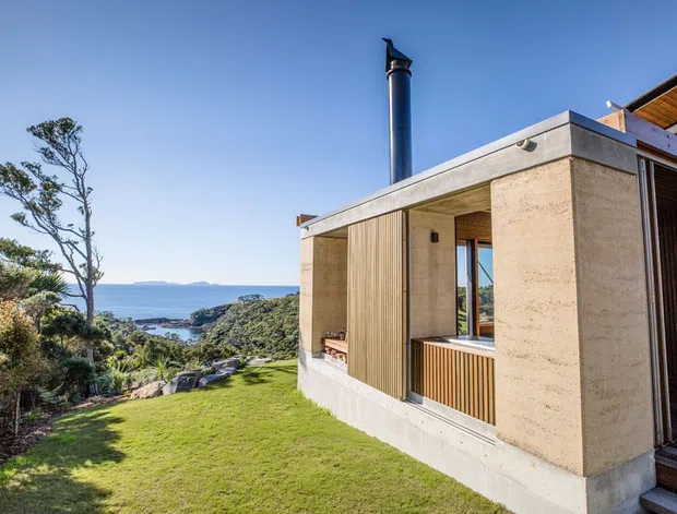 Exterior view of a contemporary coastal house set into a landscaped hillside, with a flat concrete roof, pale textured concrete walls, vertical timber screening, deep recessed openings, and a prominent dark metal chimney. The building overlooks dense coastal vegetation and the sea beyond, with a bright blue sky and lawn in the foreground.