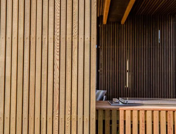Close-up architectural view of a timber-clad exterior and covered outdoor deck or balcony. The facade uses closely spaced vertical timber battens, continued across the ceiling and balustrade, with exposed timber beams and a recessed seating area.