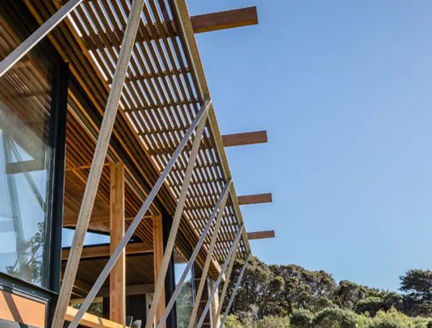 Exterior architectural photograph of a contemporary timber-clad building set within a landscaped bush setting. The facade features extensive vertical timber slatting forming a screened upper structure, exposed timber posts and beams, large glazing, diagonal bracing, projecting timber members, and a raised deck or platform. The building sits beside a manicured lawn and native-looking vegetation under a clear blue sky.