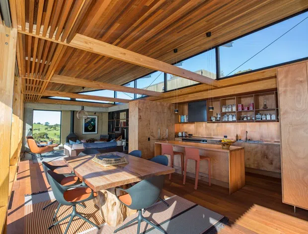 Open-plan dining and home bar area with a long timber dining table, sculptural upholstered chairs, extensive built-in timber cabinetry, and a compact drinks bar. Exposed timber beams and slatted timber ceiling create a warm architectural feature, while large glazed openings provide expansive views and abundant natural light.