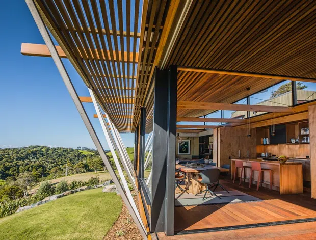 A modern timber-clad outdoor living and dining space opening onto a landscaped sloping site. The image shows extensive timber overhead louvres and soffits, exposed structural framing, dark vertical supports, large glazed openings, and a built-in timber kitchen/bar area with dining furniture. The interior connects directly to a terrace and expansive green landscape.