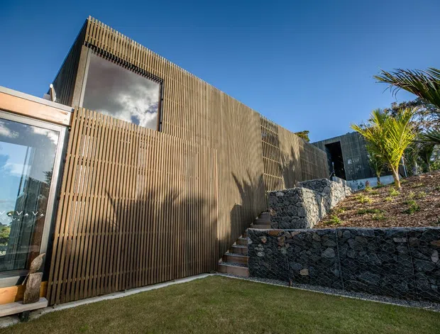 Exterior view of a contemporary, low-profile building on a landscaped sloping site. The facade is predominantly clad in closely spaced vertical timber battens, with large glazed openings and a timber-framed glazed section at left. Stone-filled gabion retaining walls and a stairway connect different garden levels, with tropical planting visible at right under a clear blue sky.