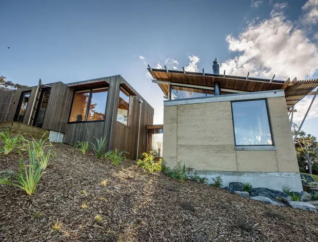 Exterior view of a modern, compact residential building on a sloped landscaped site. The structure combines dark vertical timber cladding, exposed concrete walls, large windows, and a projecting timber-framed roof canopy with a chimney. Native-style planting and mulch surround the building.