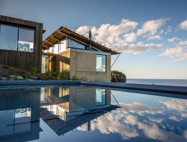 A contemporary coastal residence viewed from beside an infinity-edge swimming pool. The house features dark vertical timber cladding, exposed timber roof structure, large glazed openings, and pale stone or concrete walls. The pool reflects the building and sky, with an expansive ocean horizon beyond.