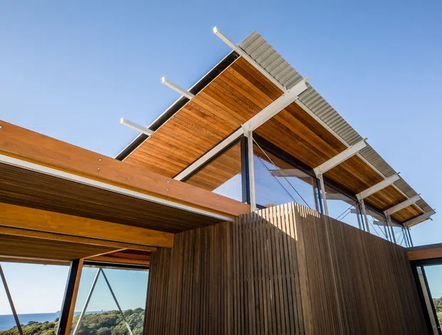Low-angle exterior view of a contemporary timber-clad building with a dramatic elevated roof canopy, exposed timber soffits, slender steel supports, and large glazed openings. The structure appears set on a coastal or elevated site, with distant landscape and water visible beyond.