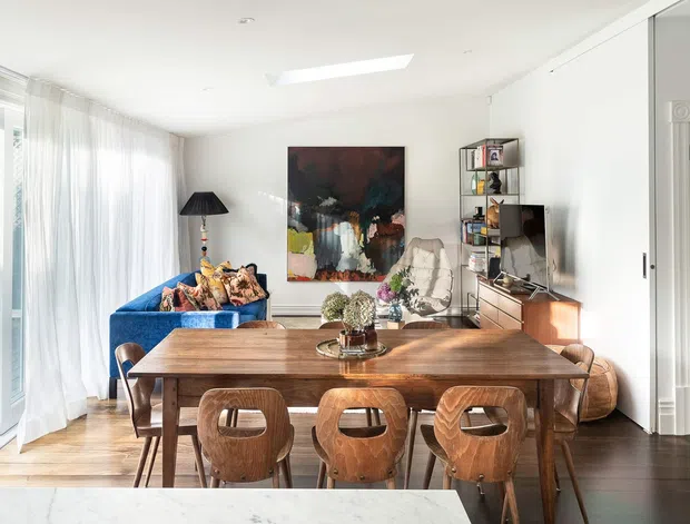 Bright open-plan dining and living area with a large rustic timber dining table and sculptural wooden chairs, a vivid blue sofa, artwork, shelving, and generous sheer-curtained glazing. The room combines warm timber finishes with a contemporary, eclectic furnishing scheme.