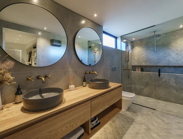 Contemporary bathroom with a double floating timber vanity, two round vessel basins, wall-mounted bronze-toned tapware, large circular mirrors, and a spacious frameless glass shower enclosure. The room features dark grey textured wall tiles, coordinated floor tiles, warm timber joinery, recessed lighting, and a high horizontal window.
