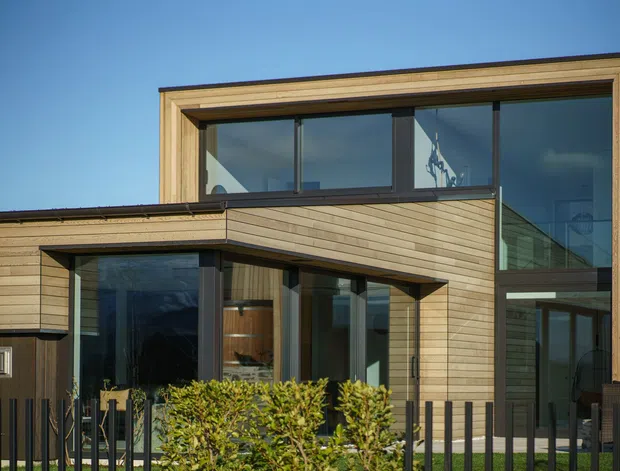 Exterior view of a contemporary two-storey home with clean rectilinear volumes, extensive dark-framed glazing, and horizontal light timber cladding. A projecting lower-level section creates a sheltered outdoor area, with a small landscaped frontage and black metal fencing.