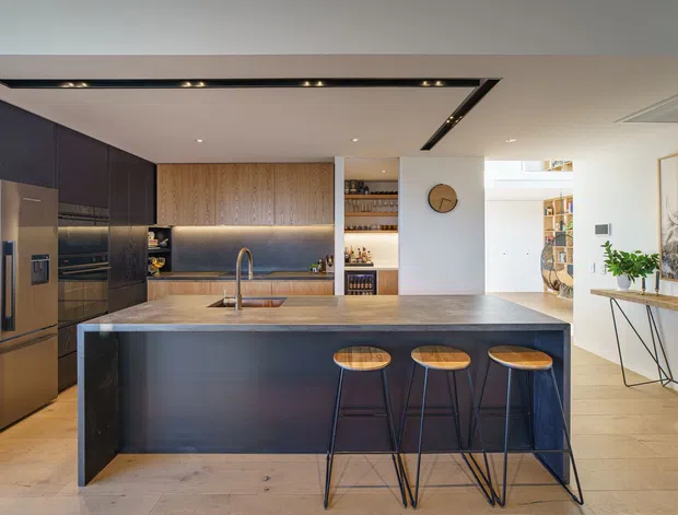 A contemporary open-plan kitchen dominated by a large dark island with three stools, integrated dark cabinetry and appliances, a timber-accented back wall, stone benchtops, open shelving, and a small adjacent dining or living area. Recessed linear lighting and warm timber flooring create a refined, modern interior.