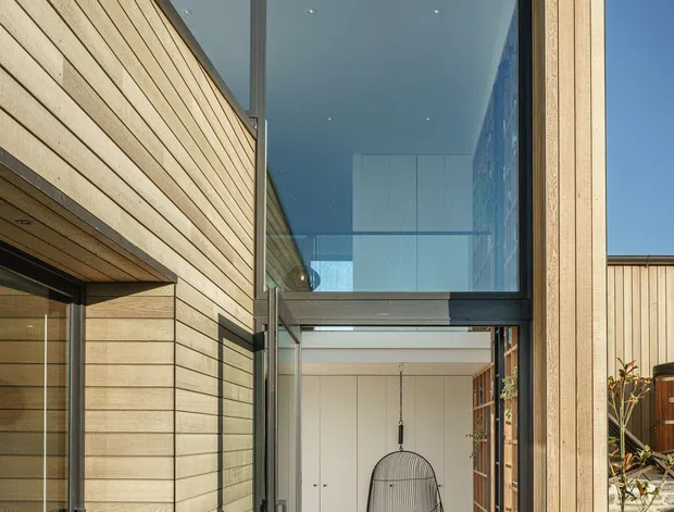A modern timber-clad residential entry courtyard with a dramatic double-height glazed opening. Pale horizontal timber boards frame the facade and soffit, while dark metal window and door frames contrast with the warm wood. Through the open glass entry, a compact interior foyer with pale cabinetry, timber flooring, and a hanging chair is visible. Gravel edging, concrete paving, and a low stone wall complete the exterior approach.