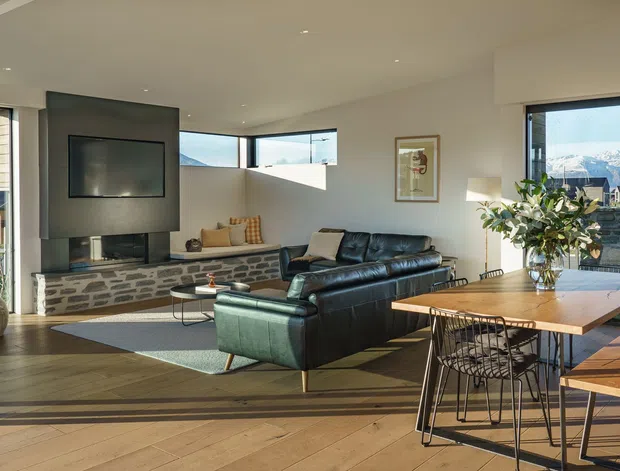 Bright open-plan living and dining area with expansive glazing and mountain views. The room features wide-plank timber flooring, a stone-faced fireplace wall with built-in television, a dark leather sectional sofa, and a large timber dining table with wire-frame chairs and bench seating. Contemporary furnishings and abundant natural light create a warm, airy atmosphere.