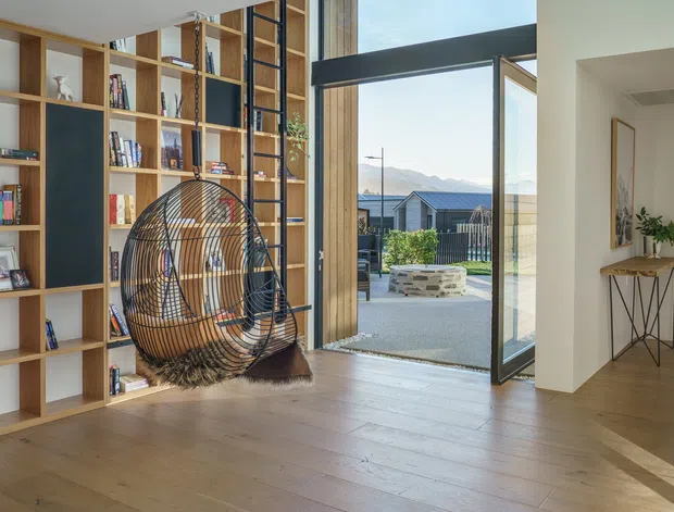 A bright, contemporary open-plan living area with extensive built-in timber shelving, integrated dark display panels, a hanging rattan egg chair, and wide timber flooring. Full-height glazing and an open glass door connect the room to an outdoor terrace, with mountain scenery visible beyond. A hallway and console table lead to another glazed living space.