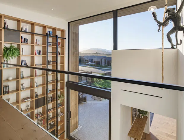 A bright, contemporary double-height interior viewed from an upper-level landing or mezzanine. Full-height black-framed glazing overlooks a landscaped residential setting and distant hills. A large built-in timber bookshelf with integrated ladder lines the left wall, while a playful suspended monkey sculpture hangs beside the stair opening. Pale timber flooring, white walls, recessed downlights, and black metal balustrades create an airy, refined architectural interior.