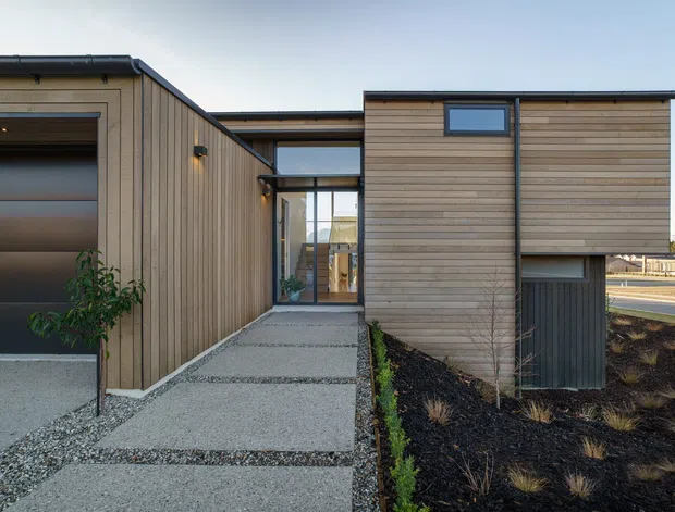 Contemporary single-storey building exterior with horizontal and vertical timber cladding, dark metal roof edges and trims, and a glazed entry connection. A textured concrete path leads between the timber-clad volumes, with a planted garden bed and roadway visible beyond.