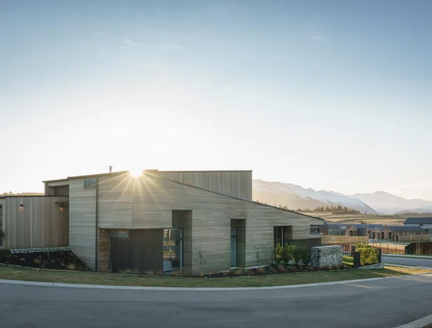 Exterior view of a contemporary single-storey house with horizontal timber cladding, a low-pitched roofline, dark-framed openings, and a simple landscaped frontage. The home sits in a suburban setting with mountains visible in the distance.