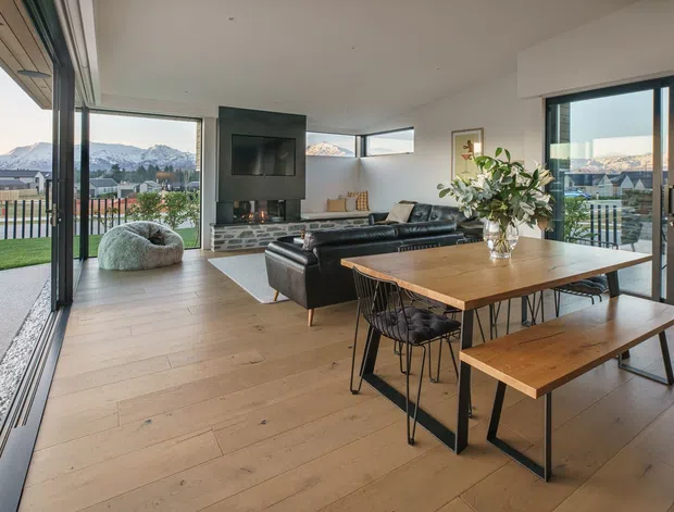 Open-plan contemporary living and dining area with full-height sliding glass doors opening to a terrace and lawn. The room features light timber flooring, a black-framed fireplace or media wall with stacked-stone detailing, a dark leather sectional sofa, and a timber dining table with bench seating. Mountain and suburban views are visible beyond the glazing.