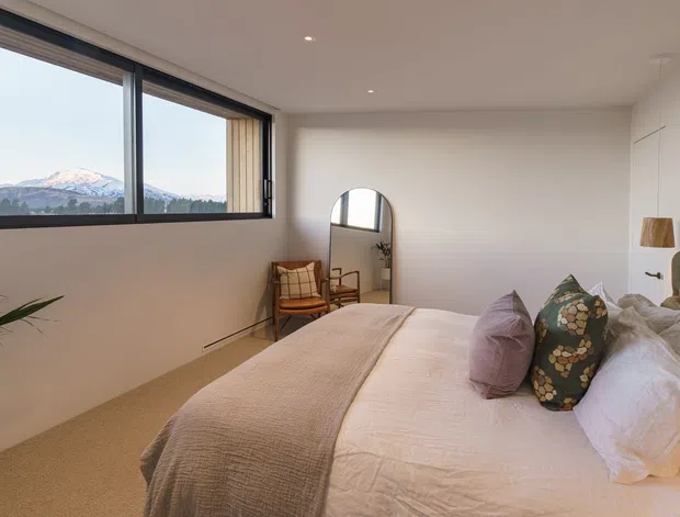 A bright, contemporary bedroom with a large corner window overlooking a mountainous landscape. The room features a low upholstered bed with layered neutral bedding and patterned cushions, a tufted green headboard, warm beige carpet, a potted plant, a timber lounge chair, and a freestanding arched mirror. Minimal recessed lighting and white walls create an airy, calm atmosphere.