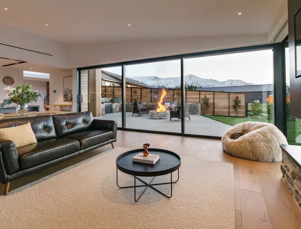 Contemporary open-plan living room with a black leather sofa, round coffee table, textured lounge chair, timber flooring and a large built-in fireplace feature wall. Floor-to-ceiling sliding glass doors open to a paved courtyard with an outdoor fire, with mountain scenery visible beyond.